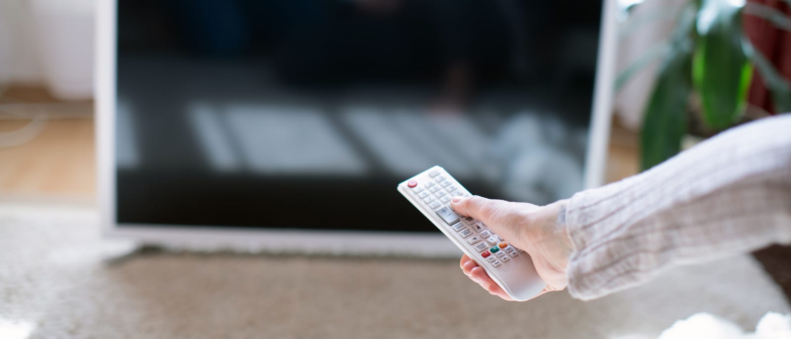A hand holding a television remote control aimed at a glowing screen in a living room