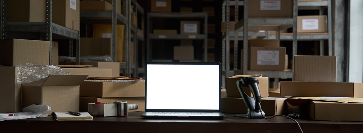 An online seller packing e-commerce orders at a laptop in a small warehouse