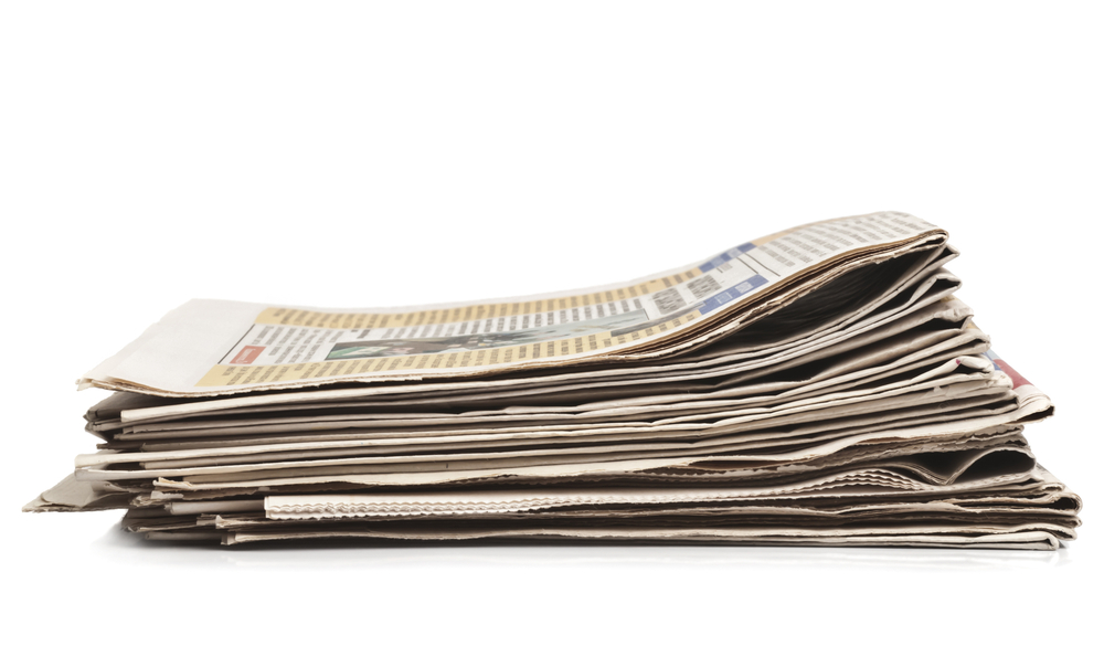 Stacks of printed newspapers beside a glowing computer screen displaying text